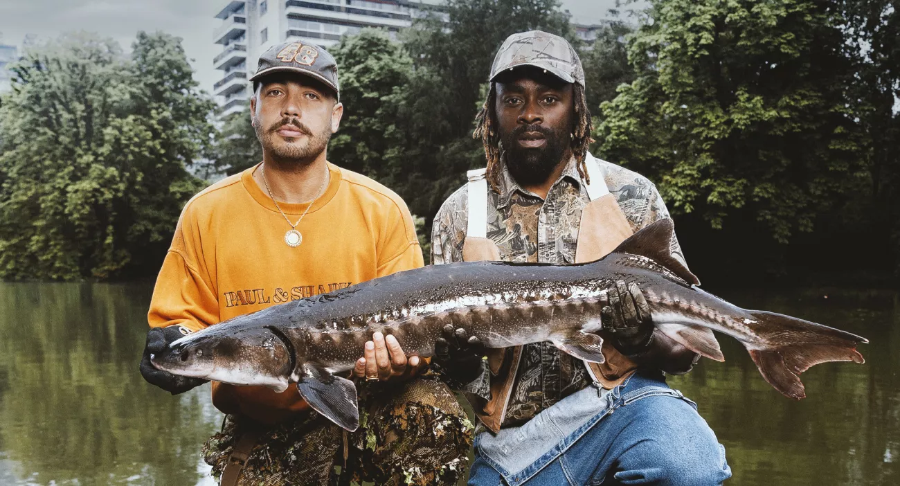 Photo de Isha et Limsa, habillés en pêcheurs, tenant en leur main un poisson. Ils sont assis devant une rivière, avec des immeubles en arrière plan. Il s'agit de l'image officielle de leur tournée.