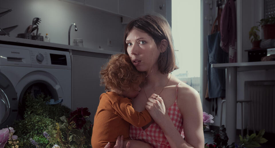 Photo d'Amandine Laval, comédienne du spectacle, tenant un enfant dans ses bras. En arrière plan une machine à laver ouverte d'où sortent et tombent sur le sol un grand nombres de fleurs.